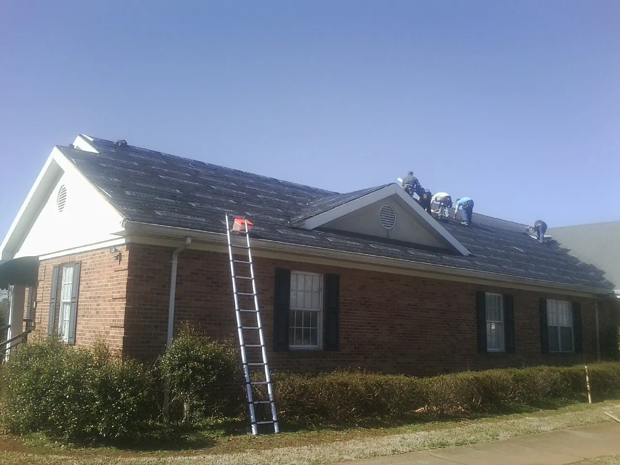 Crew tearing shingles off a brick ranch roof with a ladder set — Emergency Roof Repair work by our crew in Bowman